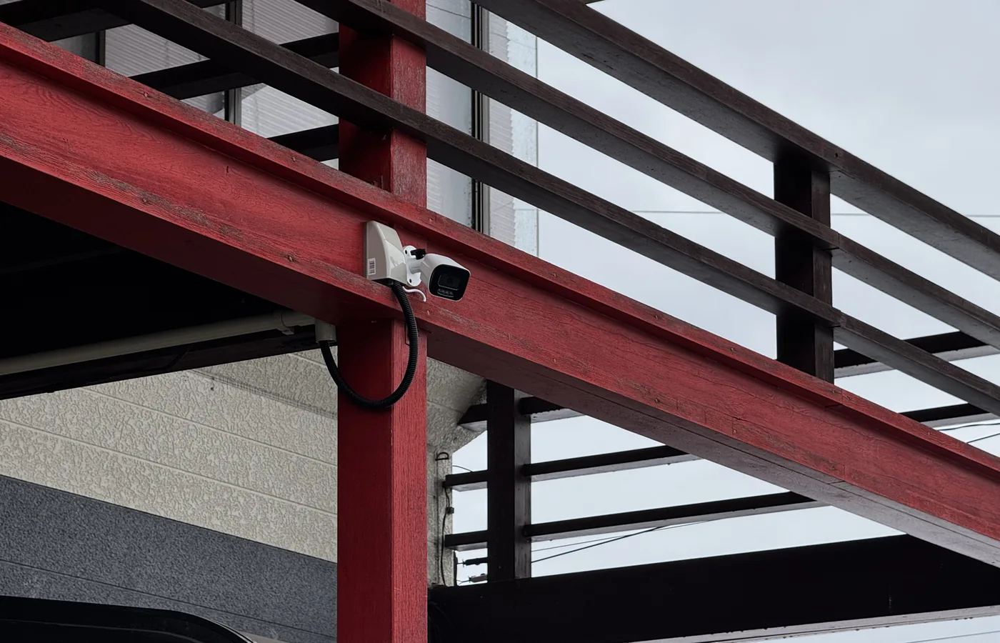 建物の赤い梁に設置した屋外用バレット型防犯カメラと、可とう管で保護された配線